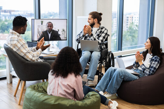 Diverse group of colleagues including man in wheelchair and a woman engaging in virtual meeting with African male speaker on screen, demonstrating teamwork, inclusivity, online collaboration.