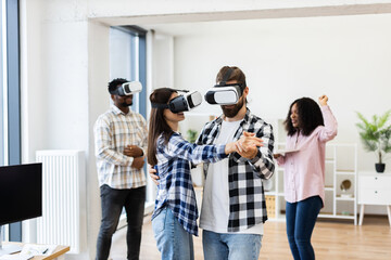 Diverse group of young professionals enjoying activities with virtual reality glasses inside office setting. Two dancing while others laugh, demonstrating happiness and creativity in technology.