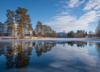 Reflection of pine trees and modern residential buildings on a calm partially frozen lake under clear blue sky in winter morning light