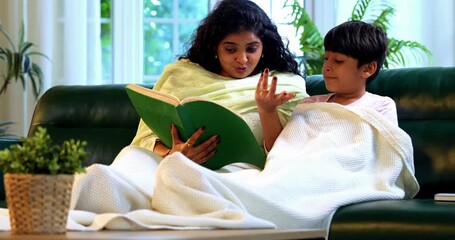 Indian mother son reading a story book together while sitting on sofa in modern home, wrapped in cozy shawl, sharing bonding time, warmth, affection, parent child relationship, lifestyle indoors - Powered by Adobe
