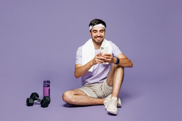 Full body young strong fitness trainer instructor sporty man sportsman wear sportswear train in gym sit hold use mobile cell phone isolated on plain purple background. Workout sport fit abs concept.