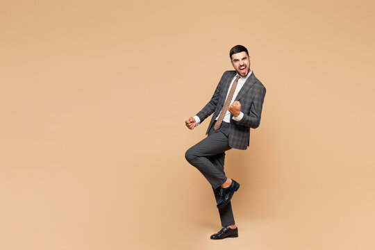 Full body young happy successful employee business man corporate lawyer wear classic formal grey suit shirt tie work in office do winner gesture raise up leg isolated on plain beige background studio.