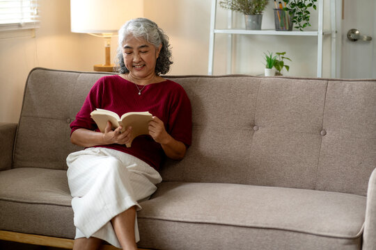 Asian older adult woman relaxing reading book on sofa home living room enjoying peaceful cozy indoor lifestyle education and personal growth