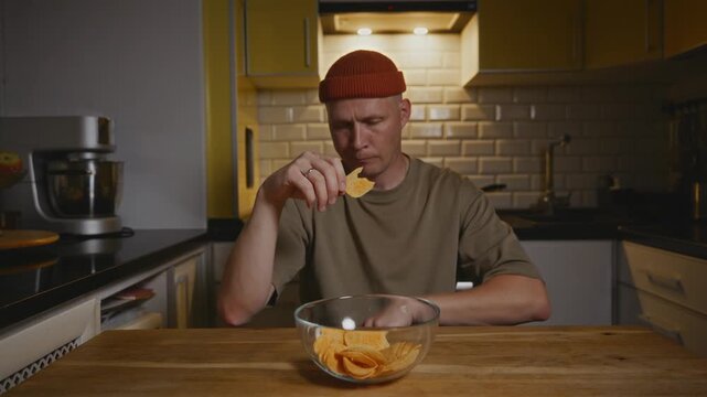 A man sits at the kitchen table in the evening, calmly eating chips from a glass bowl. Warm light creates a cozy atmosphere of mindful snacking and evening rituals. High quality 4k footage