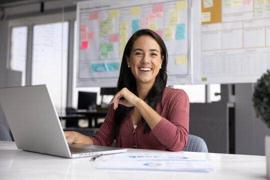 Businesswoman sit at desk, smile at camera while using laptop