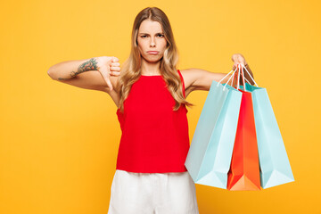 Young sad woman wear red casual clothes hold in hand paper package bags after shopping showing thumb down dislike gesture isolated on plain orange yellow background. Black Friday sale buy day concept.
