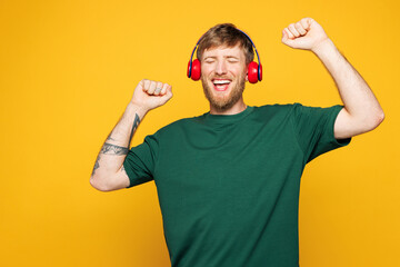 Young smiling happy Caucasian man he wear green t-shirt casual clothes listen to music in headphones raise up hands dance isolated on plain yellow orange background studio portrait. Lifestyle concept.