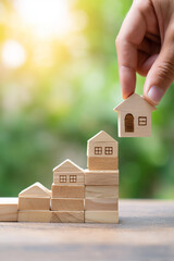 Wooden blocks depict a rising housing market, with a hand placing the final piece of the house on top, symbolizing achievement and investment.
