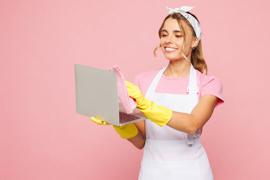 Young IT woman she wear white apron hold basin with detergent bottles while doing housework tidy up wiping laptop pc computer screen with cloth isolated on plain pink background. Housekeeping concept.