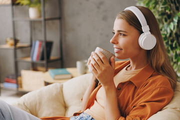 Close up young woman wear orange casual clothes listen music in headphones drink coffee sits in chair stay at home hotel flat rest relax spend free time in living room indoor. People lounge concept.