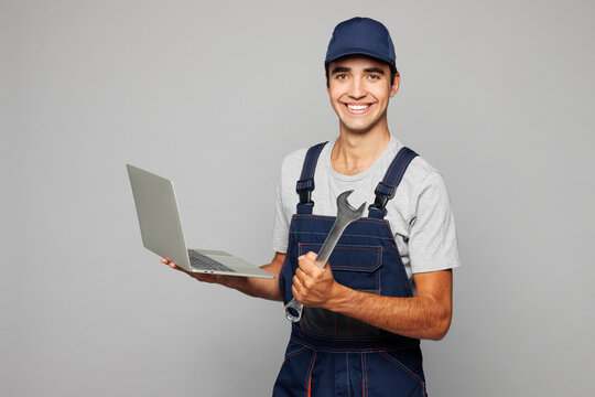 Young mechanic IT man wearing overall hat uniform workwear clothes work in garage using laptop pc computer to diagnose car hold wrench isolated on plain grey background. Automotive repair job concept.