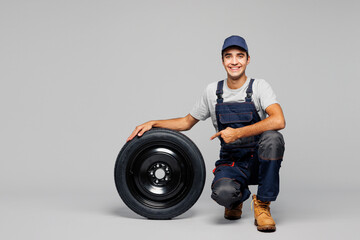 Full body young smiling happy car mechanic man wear overall hat uniform workwear clothes work in garage sit point index finger on wheel isolated on plain grey background Automotive repair job concept.
