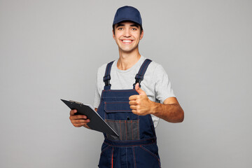 Young car mechanic man wear overall uniform workwear clothes work in garage hold clipboard with paper account documents show thumb up isolated on plain grey background. Automotive repair job concept.