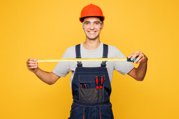 Young smiling employee laborer handyman man he wear helmet overall hold measuring tape isolated on...