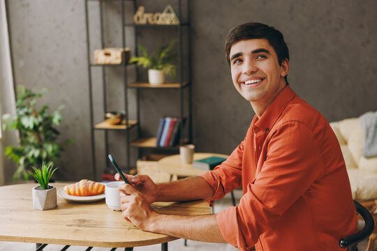 Side view young man wearing orange casual clothes use cell phone sitting alone at table in coffee shop cafe relax rest in restaurant during free time indoors. Freelance mobile office business concept.