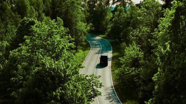 Van driving on forest road. Black minivan moving along curvy highway surrounded by dense trees on sunny afternoon. Vehicle traveling through winding green landscape