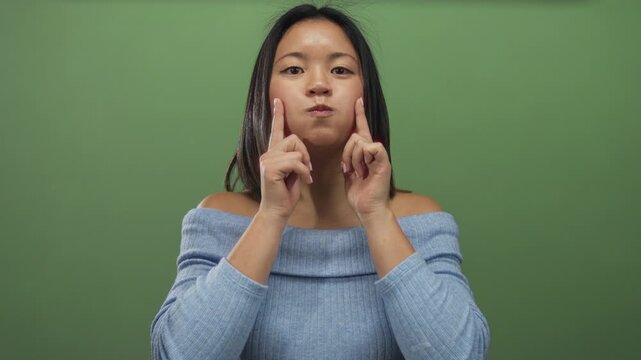 Woman in blue sweater with puffed cheeks poses against a plain green wall demonstrating a playful expression isolated in a studio setting.