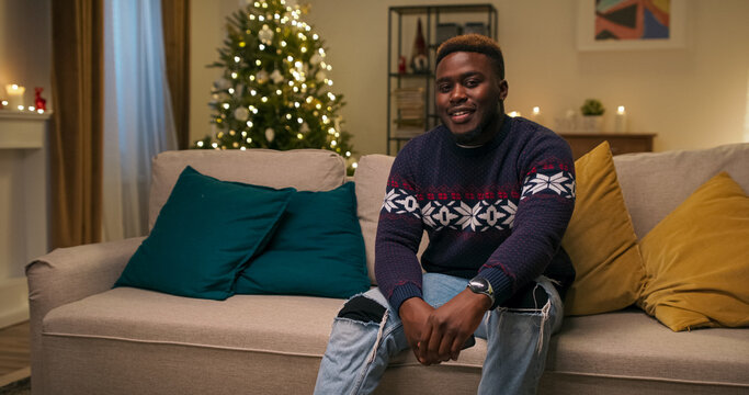 Attractive young guy sitting on the couch. The guy looks at the camera and smiles. He is wearing warm holiday sweater, ripped jeans and watch. A Christmas tree flickers behind.