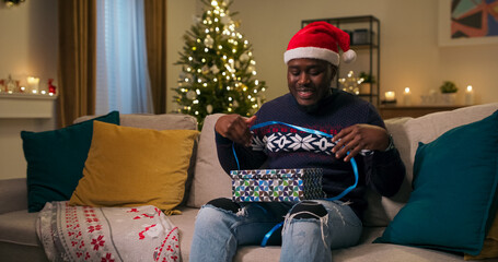 A young man of multi-racial background in a Santa Claus hat sits on a sofa on Christmas Eve, unpacking gifts. He opens the box, finds warm headphones he doesn't need, and, upset, puts them aside.