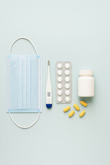 Flat lay medical objects with face mask, digital thermometer, pills and medicine bottle on pastel background