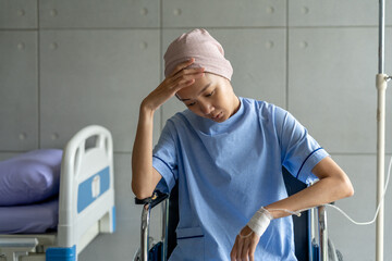 A cancer patient sits in a wheelchair with an IV drip looking stressed and sad. Asian Woman receiving IV therapy in a wheelchair showing visible signs of stress and depression
