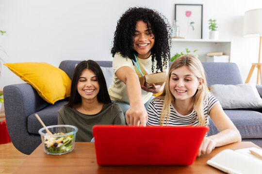 Three cheerful multi-ethnic young women using laptop while eating takeaway food in living room. Female friendship and youth concept