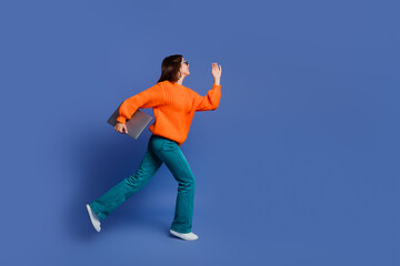 Young woman with orange sweater and laptop walking against a blue background highlighting fashion...