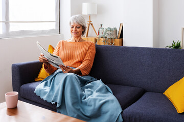 Smiling senior retired woman wrapped in a warm blanket reading newspaper on sofa at home in winter season