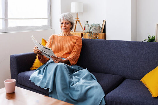 Smiling senior retired woman wrapped in a warm blanket reading newspaper on sofa at home in winter season