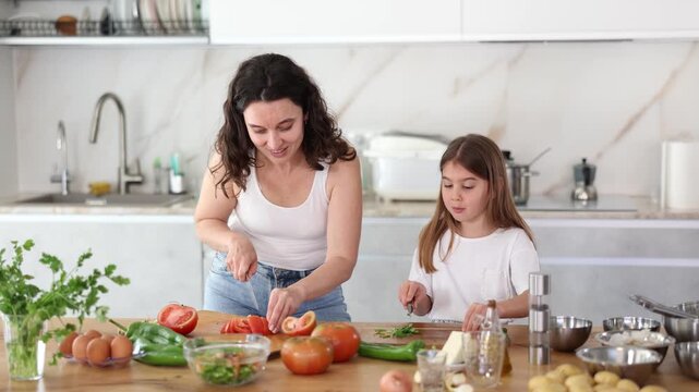 Happy mother and her little daughter are cutting fresh vegetables for salad in the kitchen - Powered by Adobe