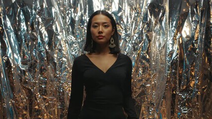 Camera zooming in on Asian model standing against foil backdrop in studio, revealing earrings - Powered by Adobe