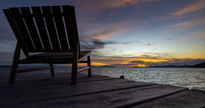 Atmospheric time lapse video of a sunset over a tranquil ocean at Papua, Indonesia, with a lone wooden chair resting on a pier. The warm hues of the sky reflect on the water as the light gently fades 
