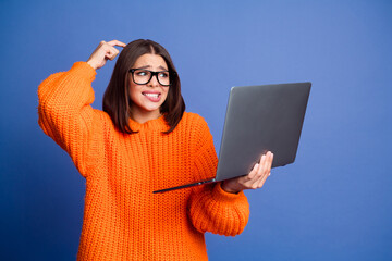 Confused young woman wearing orange sweater using a laptop against purple background, showcasing an...