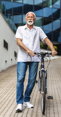 Happy senior man walks his bicycle outdoors and smiles. Cheerful old healthy man enjoy a peaceful walk with bike in a urban scene. Elderly man in casual outfit strolls through the city with bicycle.