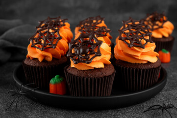 Halloween chocolate cupcakes with orange frosting and chocolate spider web on a dark background. Festive dessert. Funny food idea. Copy space.