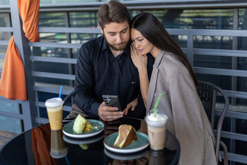 A couple is sitting at a table in a cafe, looking at a smartphone, with drinks 