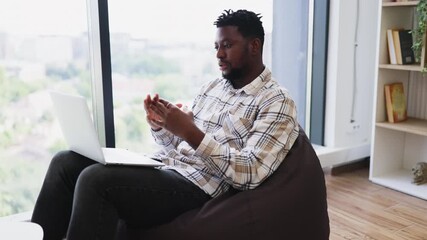 Young adult African American man sitting on bean bag using laptop for video call, communicating confidently in modern room with bookshelf and window view, showcasing work from home