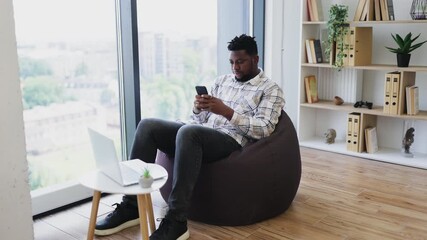 Adult man relaxing on bean bag chair inside modern room working on smartphone near laptop. - Powered by Adobe