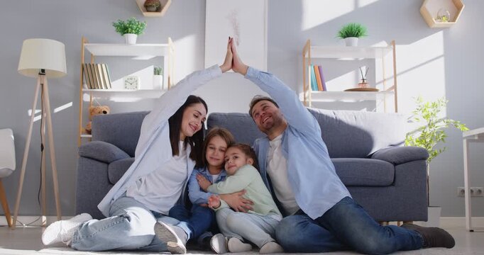Happy parents and children at home. Smiling mother and father make symbolic house roof with arms over kids while sitting on floor by couch in living room. Home, family, love, care, protection concept