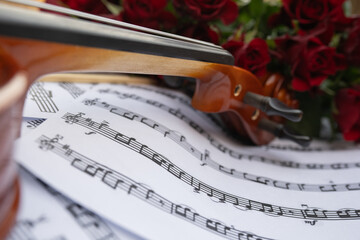 Close-up of a wooden violin resting on sheet music surrounded by vibrant red roses, creating a...
