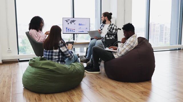 Group of young adults, diverse gender and race, working together in modern casual office. Collaborative atmosphere, bean bags, wheelchair accessibility, and digital devices convey teamwork, inclusion.