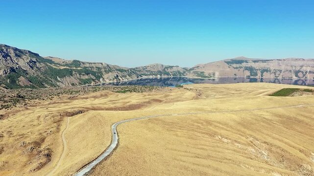 Small car drives across golden plateau at Nemrut Crater, winding road above blue caldera lake, wide summer landscape &mdash; concepts: freedom, journey, minimalism, solitude, exploration, escape