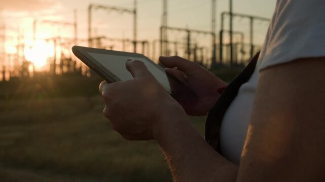 Electrical engineer's hands typing on tablet against background of high voltage transformer substation in evening at sunset