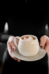 Woman holding a white mug of hot chocolate with marshmallows. Cozy winter mood, black nails, dark background. Warm drink and comfort concept, close-up.