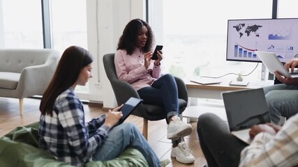 Two female colleagues, one brunette and one curly-haired, using smartphones while sitting in modern office space during informal discussion. Scene conveys teamwork, communication, technology use - Powered by Adobe