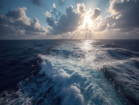 A ship's wake cuts through blue ocean under a partly cloudy sky with sunbeams