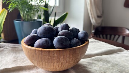 A wooden bowl filled with ripe plums sits on a light-colored fabric, with indoor plants and wooden coat hangers in the background.