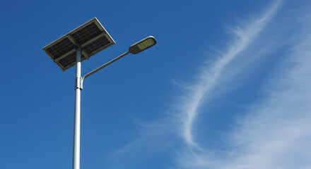 A tall solar-powered street light stands against a bright blue sky with wispy clouds.