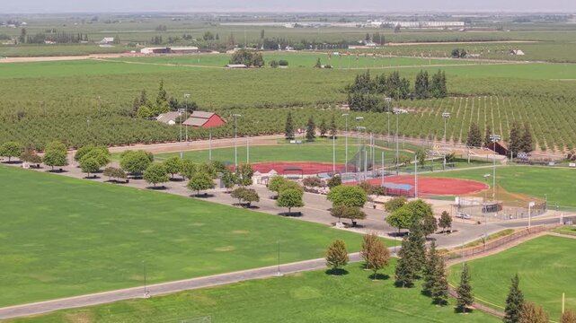 Side aerial shot over baseball fields with parking trees, farmland, road, and houses in Ripon California.