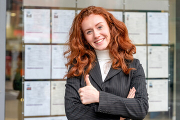 A cheerful woman, a real estate agent, is giving a thumbs up next to a window showcasing property listings